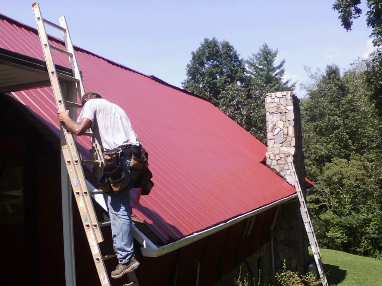 Roofer on a ladder beside a red metal roof and stone chimney — Soffit & Fascia Repair by Roofing Brothers in Primm Springs